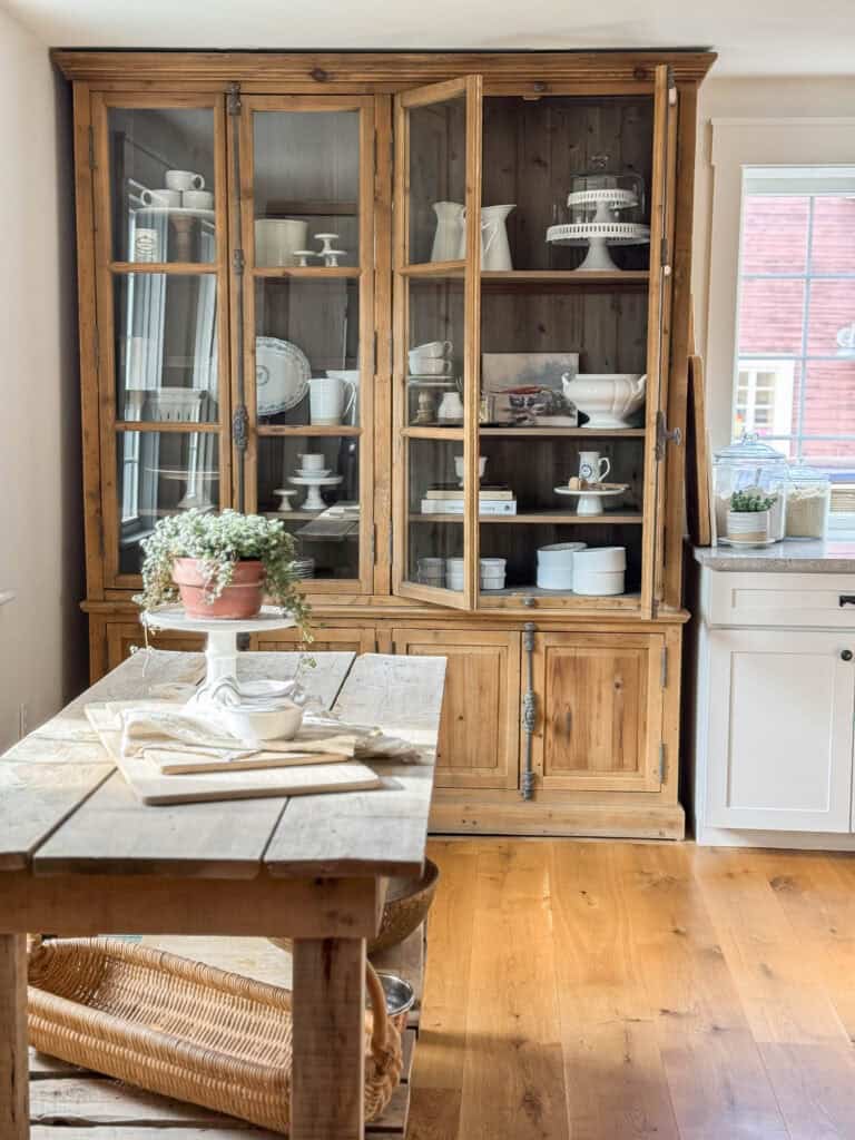 Farmhouse kitchen nook with rustic wooden prep table centered in front of a large glass-front hutch filled with white dishes, natural wood floors, and soft window light creating a functional small space layout.