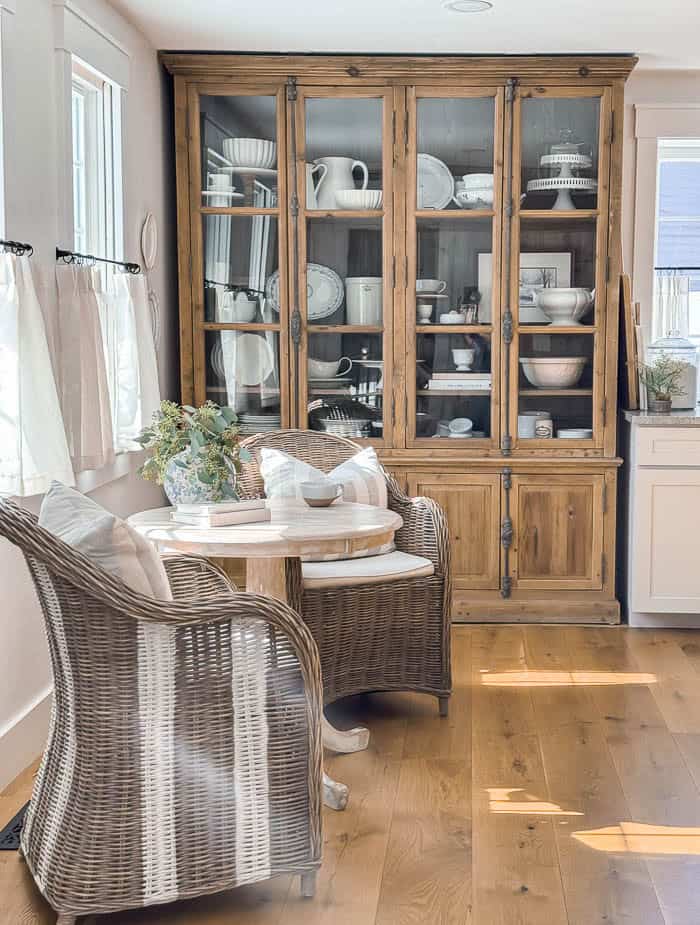 Cozy kitchen nook seating area with wicker chairs and a small round table styled in front of a glass cabinet, natural light streaming through windows in a warm farmhouse kitchen setting.