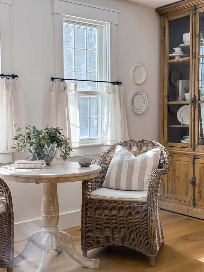 Cozy kitchen nook seating area with wicker chairs and a small round table styled in front of a glass cabinet, natural light streaming through windows in a warm farmhouse kitchen setting.