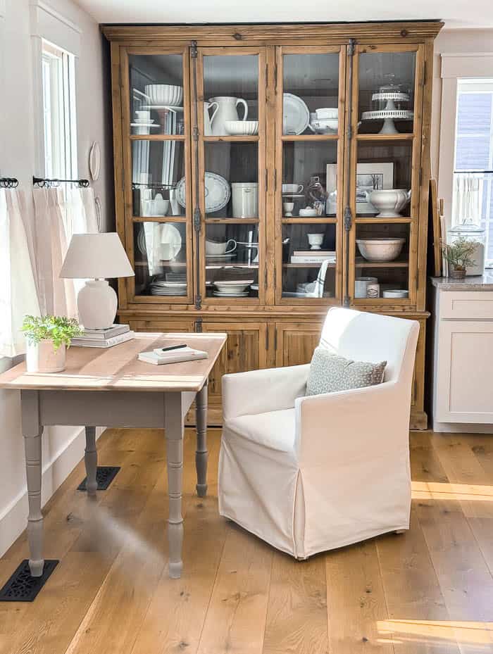 Small farmhouse kitchen nook styled as a home office with a wooden desk, white slipcovered chair, table lamp, and large hutch backdrop, demonstrating a practical small space design idea.