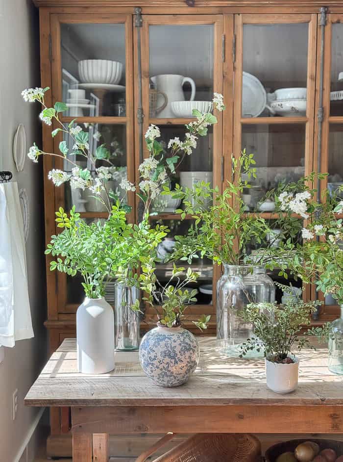 Realistic faux greenery stems and faux flowering branches arranged in glass and ceramic vases for spring decorating.