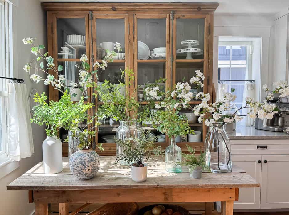 Realistic faux greenery stems and faux flowering branches arranged in glass and ceramic vases for spring decorating.
