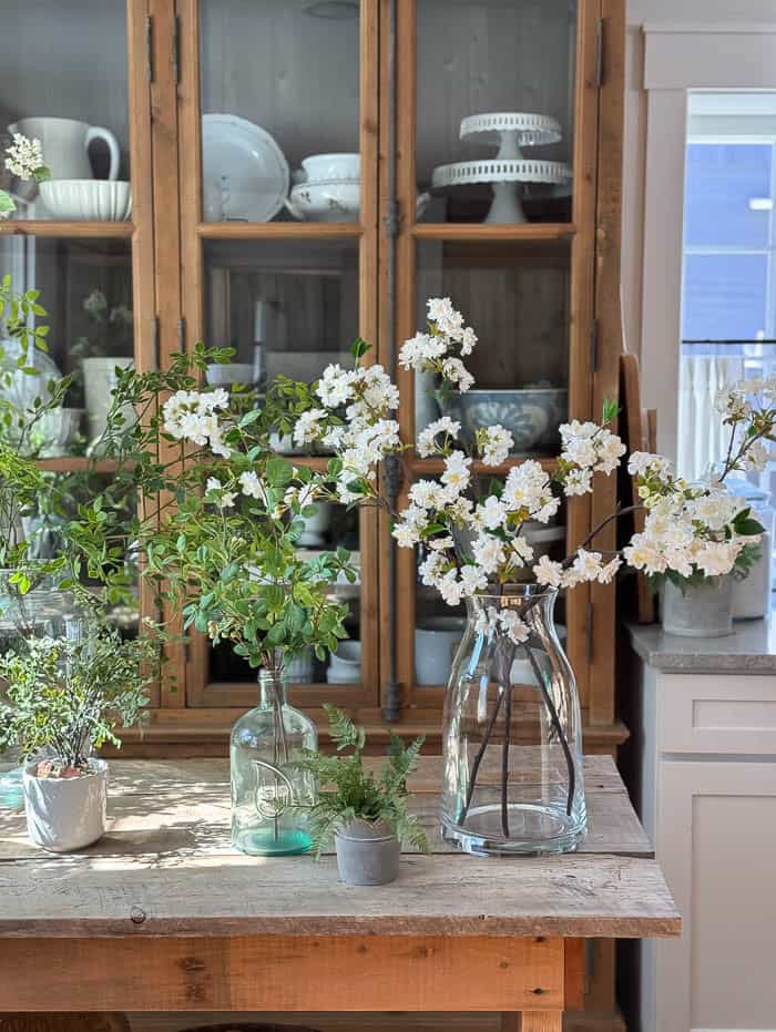 Collection of realistic faux stems and greenery styled in glass vases on a rustic kitchen island for spring decorating.
