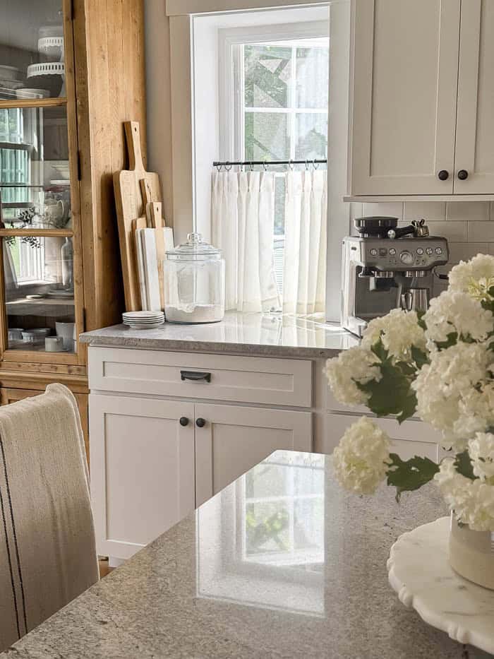 Close-up of simple linen café curtains mounted on black curtain rods in a cottage-inspired kitchen filled with natural light and soft neutral tones.