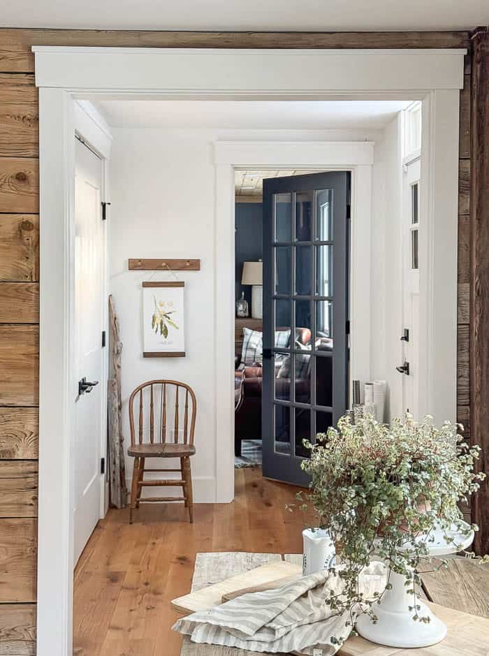 Front hallway before wallpaper, with white walls, wood floors, and a wooden chair beside a dark glass-paneled door