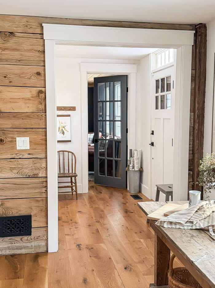 Front hallway before wallpaper, with white walls, wood floors, and a wooden chair beside a dark glass-paneled door, a white front door with transom windows
