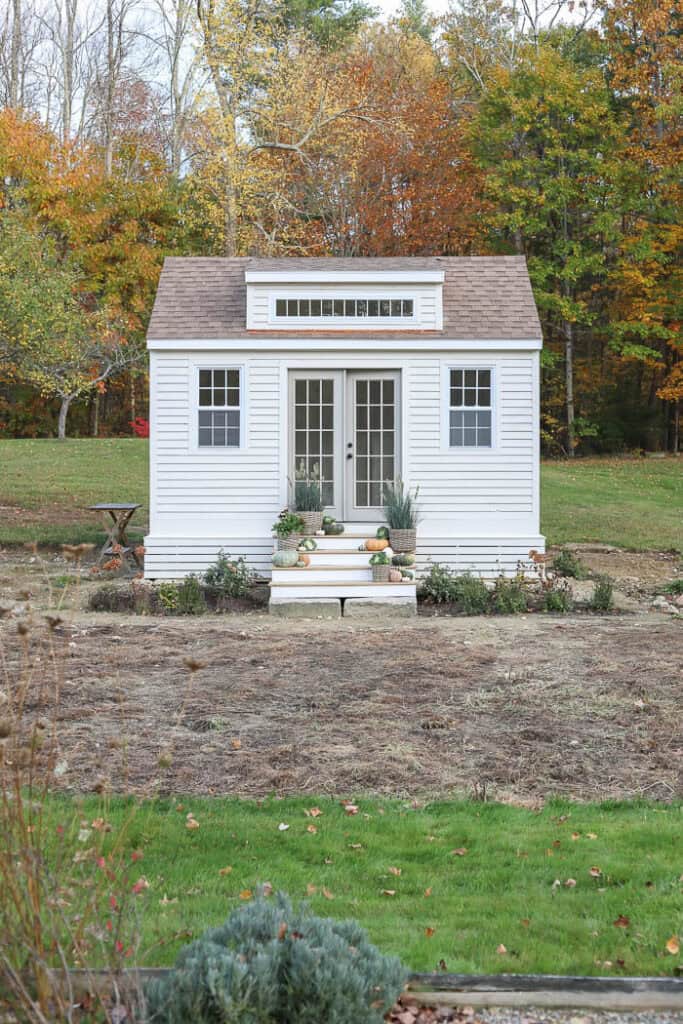White cottage-style garden shed with French doors surrounded by autumn foliage