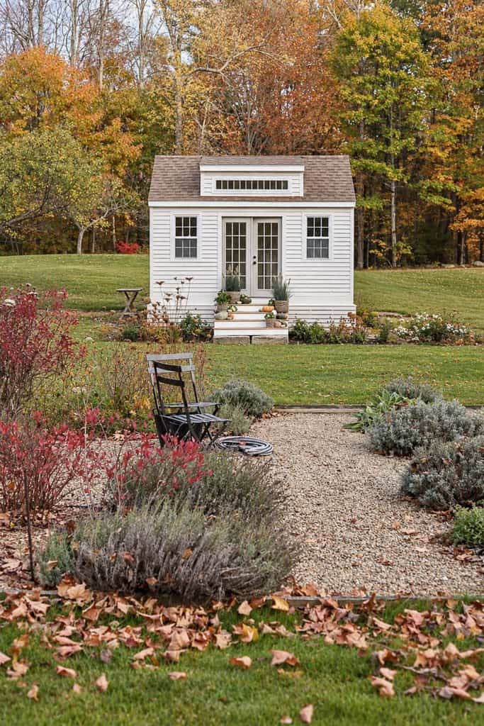 White cottage-style garden shed with French doors surrounded by autumn foliage