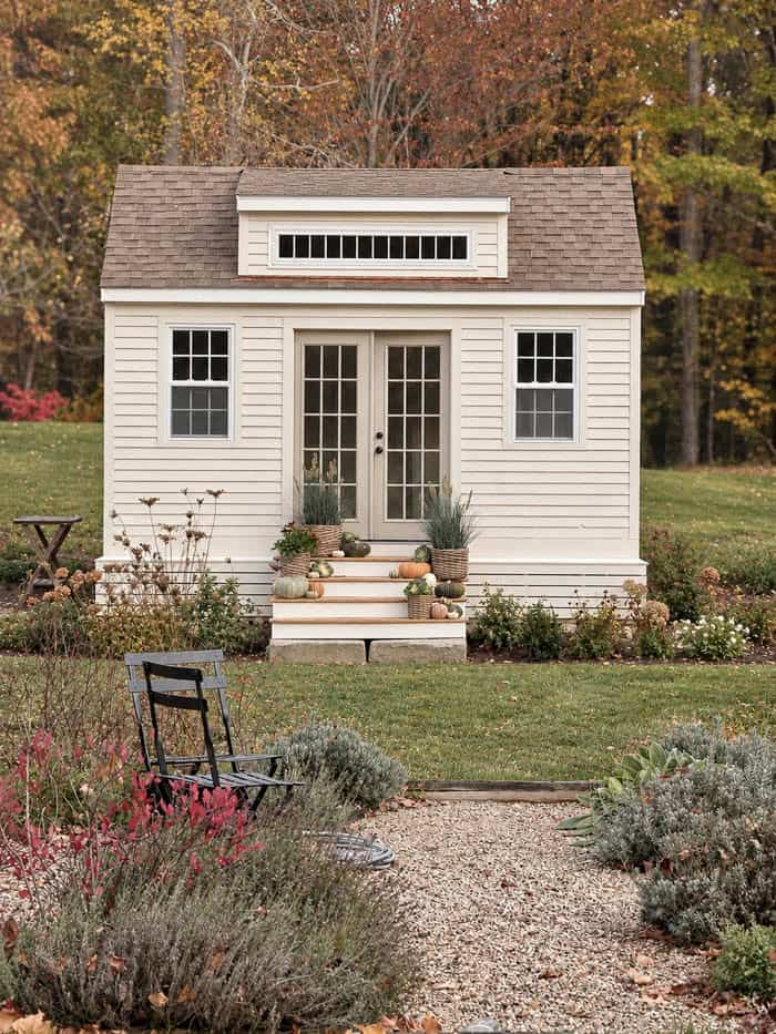 White cottage-style garden shed with French doors surrounded by autumn foliage